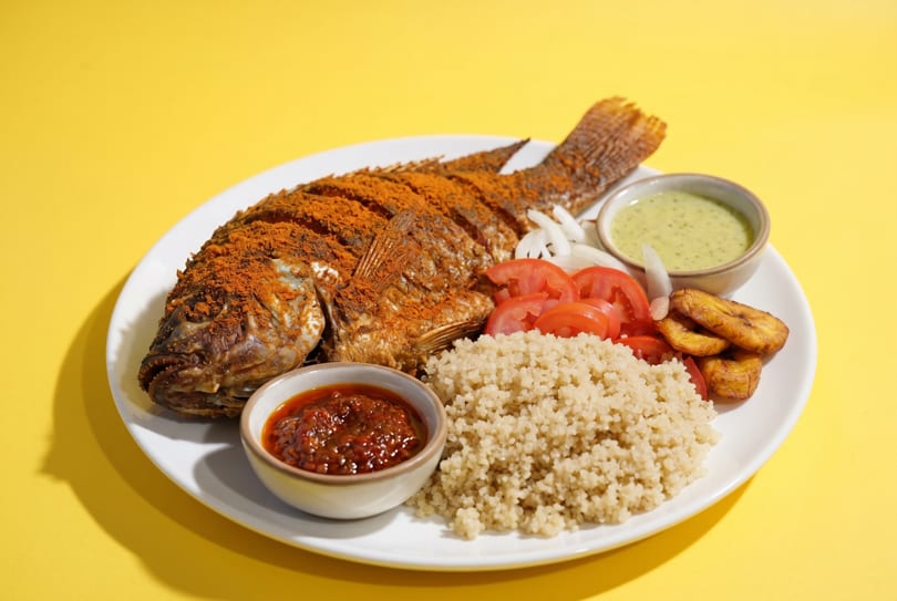 Whole fried fish with red spice, quinoa, sliced tomatoes, onions, plantains, and two sauces.