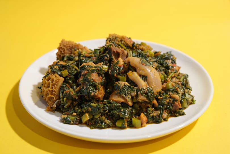 A white plate holds a vibrant green stew with meat and tripe against a bright yellow background.